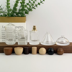 Op de afbeelding: Een verzameling heldere glazen parfumflesjes en houten stoppers. De flesjes hebben verschillende vormen, waaronder rechthoekige, vierkante en cilindrische ontwerpen. De stoppers zijn in verschillende vormen en kleuren, waaronder bruin, zwart en natuurlijk hout.