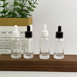 May include: Four clear glass dropper bottles with white and black caps are arranged on a wooden surface. The bottles are empty, and a green plant is visible in the background. The bottles are for essential oils or cosmetic products.