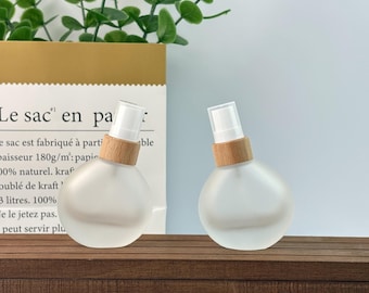 May include: Two frosted glass spray bottles with wooden collars and white spray nozzles. The bottles are round and sit on a dark wooden surface. The background includes a paper bag with French text and green leafy sprigs.
