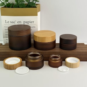 Op de afbeelding: Een verzameling bruine glazen potten met houten deksels. De potten variëren in grootte, met sommige deksels van lichtgekleurd hout en andere van donkerder hout. Witte plastic deksels zijn ook zichtbaar. De potten zijn gerangschikt op een houten oppervlak.