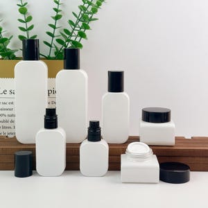 Op de afbeelding: Een verzameling witte cosmetische verpakkingen met zwarte deksels, waaronder verschillende hoge ovale flessen, twee kleinere flessen met pompdoseerders en twee vierkante potjes, waarvan er één zonder het zwarte deksel wordt getoond.