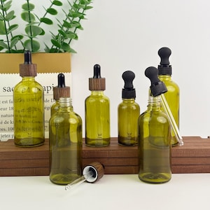 以下が含まれることがあります： 木と黒のキャップが付いたオリーブグリーンのガラス製スポイトボトルがいくつか木製の表面に配置されています。 1つのボトルはスポイトが取り外されています。 ボトルはサイズと形状が異なり、透明なガラス製スポイトがいくつか見えます。