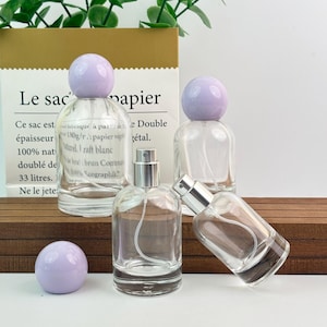 Puede incluir: Frascos de perfume de vidrio transparente con pulverizadores plateados y tapones esféricos de color lila claro. Los frascos están dispuestos sobre una superficie de madera. La imagen incluye una bolsa de papel con texto en francés.