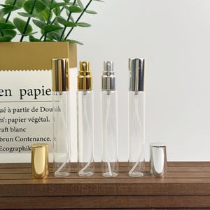 May include: Four clear glass perfume bottles with gold and silver spray tops and caps. The bottles are arranged in a row on a wooden surface. One gold cap is off to the side. The background includes a paper bag and greenery.