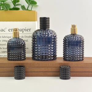 Op de afbeelding: Drie blauwe glazen parfumflessen met getextureerde diamantpatronen. De flessen hebben zwarte en gouden accenten. Twee zwarte doppen liggen voor de flessen. De flessen staan op een houten oppervlak.