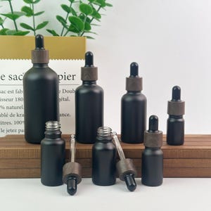 May include: A collection of matte black glass dropper bottles in various sizes, each featuring a dark wood-toned collar and a black rubber dropper bulb. Some bottles are capped, while others have their glass droppers removed and placed nearby.