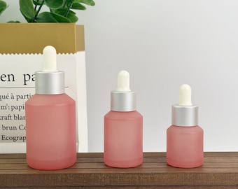 May include: Three pink frosted glass dropper bottles with silver collars and white droppers are arranged on a wooden surface. The bottles vary in size, with the largest on the left and the smallest on the right. A paper bag and greenery are in the background.