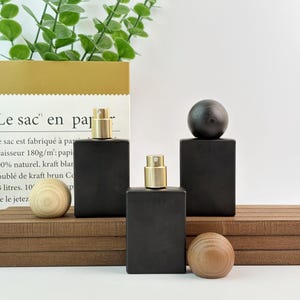 May include: Three black rectangular perfume bottles with gold spray tops and wooden spheres. The bottles are arranged on a wooden surface with a paper bag and greenery in the background.