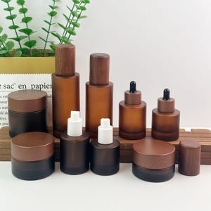 Op de afbeelding: Een verzameling amberkleurige, matglazen containers met houten deksels, waaronder potten en flessen met pomp- of druppelaars. De set bevat verschillende maten en vormen, allemaal voorzien van bijpassende donkerbruine houten doppen.