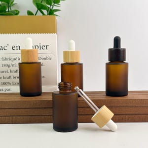 May include: Amber glass dropper bottles with wooden and black caps. One bottle is open, with the glass dropper and white tip visible. The bottles are arranged on a wooden surface, with a paper background.