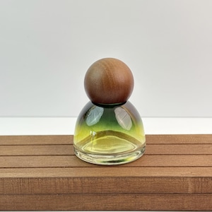 May include: A small, clear glass bottle with a rounded base and a gradient of yellow and green hues. The bottle is topped with a round, wooden stopper. The bottle sits on a brown, wooden surface.