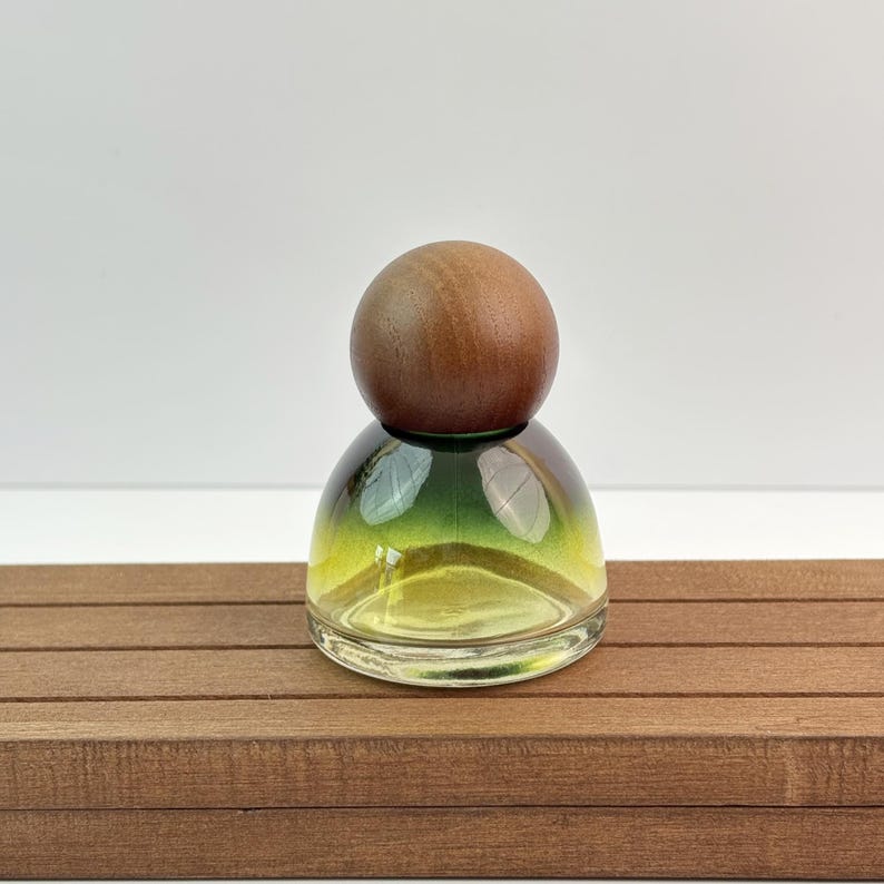 May include: A small, clear glass bottle with a rounded base and a gradient of yellow and green hues. The bottle is topped with a round, wooden stopper. The bottle sits on a brown, wooden surface.