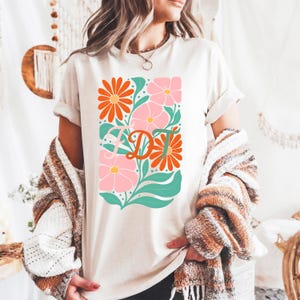 Puede incluir: Camiseta color crema con un diseño floral con flores naranjas y rosas, hojas turquesas y las letras "DT" en naranja. La camiseta tiene las mangas enrolladas y se lleva con un suéter estampado.