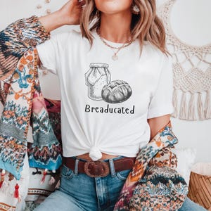 Puede incluir: Camiseta blanca con una ilustración en blanco y negro de un frasco de masa madre y una hogaza de pan. El texto "BREADUCATED" está impreso debajo de la ilustración.