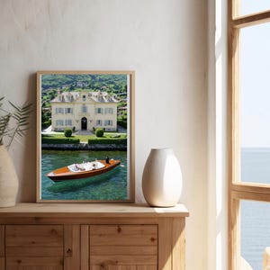 May include: A framed print depicting a lake scene with a wooden boat on the water in front of a large white building. The boat has a dark brown hull and is surrounded by clear water. The building has a green lawn and trees in the background.