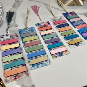 May include: Five rectangular clear acrylic bookmarks with floral patterns, each featuring a colorful tassel and multiple rows of adhesive page flags. The bookmarks are labeled with text including Your Romantasy Annotating Bestie and Your Nonfiction Annotating Bestie, with various categories printed below the sticky tabs.