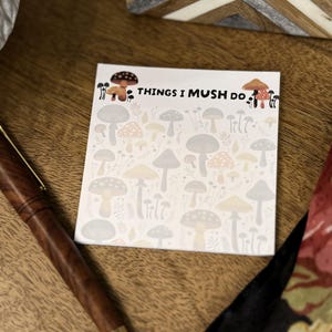 May include: A square notepad featuring a repeating pattern of various mushrooms in muted tones of gray, beige, and soft orange. The top of the notepad displays the text THINGS I MUSH DO in black lettering, flanked by two small mushroom illustrations.