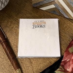 May include: A square notepad encased in clear plastic featuring a white background with a subtle light gray botanical pattern. The text "IT'S NOT HOARDING IF IT'S BOOKS" is printed in dark gray and gold lettering at the top center of the notepad.