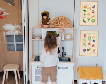May include: A child plays in a toy kitchen with a doll and a rainbow arch. Two educational posters hang on the wall, one for fruits and one for vegetables. The posters feature colorful illustrations of different fruits and vegetables.