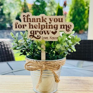 May include: A small galvanized metal bucket with faux greenery and a wooden sign that reads "thank you for helping me grow". The sign also includes the text "Love, Kasor". The bucket is tied with twine.