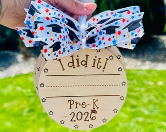 May include: Wooden graduation ornament with the text "I did it!" and "Pre-K 2026". The ornament has a decorative ribbon with graduation caps and stars. The ornament is round and has a space for writing.
