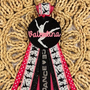 May include: A black and pink dance themed hair bow with a black circle with glitter and a white silhouette of a dancer. The circle has the name "Valentina" written in pink letters. The bow has black and pink ribbon with white polka dots and a black ribbon with white silhouettes of dancers.