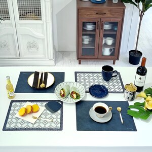 May include: A table setting with various items, including plates, cutlery, a cup of coffee, and a bottle of wine. The setting includes blue placemats, a white plate with a black napkin, and a bottle of a yellow beverage.