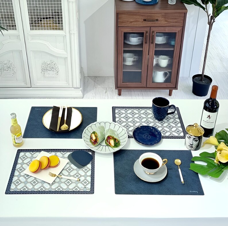 May include: A table setting with various items, including plates, cutlery, a cup of coffee, and a bottle of wine. The setting includes blue placemats, a white plate with a black napkin, and a bottle of a yellow beverage.