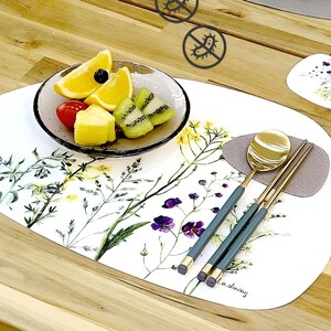 May include: Oval white placemat with a floral design, a glass bowl of fruit, and gold-coloured spoon and chopsticks. The placemat has a botanical print with yellow and purple flowers. The chopsticks and spoon have teal handles.
