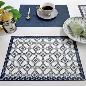 May include: A white and navy blue patterned placemat with a decorative border. A cup of coffee sits on a blue placemat, next to a decorative mug and a bowl with green food. The placemat has the text "el.a.ming".