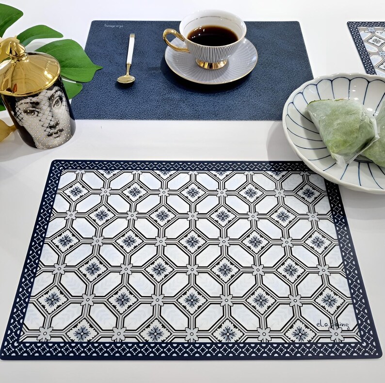 May include: A white and navy blue patterned placemat with a decorative border. A cup of coffee sits on a blue placemat, next to a decorative mug and a bowl with green food. The placemat has the text "el.a.ming".