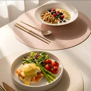 以下が含まれることがあります： ピンク色のプレースマットの上に、2つのお皿に盛られた料理が写っています。上のお皿にはグラノーラ、ベリー、バナナのスライスが乗っています。下のお皿にはエッグベネディクト、アスパラガス、トマトが乗っています。箸とスプーンも写っています。