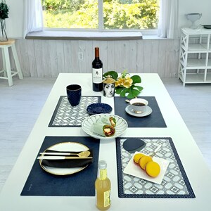 May include: A white table set for a meal with a bottle of red wine, a decorative jar, a cup of coffee, and a plate of food. The table is set with placemats, plates, and cutlery. A bottle of yellow liquid is on the table.