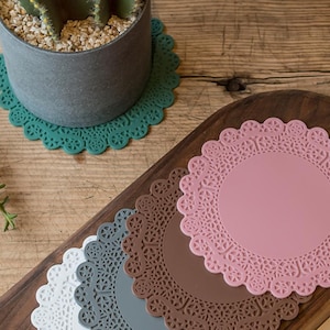 May include: A collection of decorative, round coasters in various colors, including pink, white, gray, brown, and teal. The coasters have a lace-like, scalloped edge design. A potted cactus sits on a teal coaster.