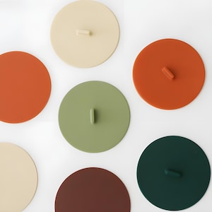 May include: A collection of colorful, round silicone lids in various shades, including orange, green, beige, and brown. Each lid features a small, rectangular handle for easy lifting. The lids are designed for covering containers.