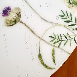 May include: A close-up of a white surface with a watercolor floral design. The design features a purple and white thistle flower, green leaves, and water droplets. The background is a light wood.