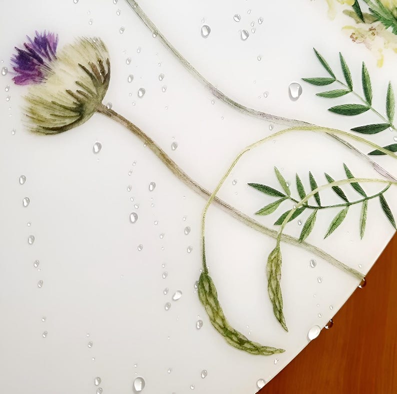 May include: A close-up of a white surface with a watercolor floral design. The design features a purple and white thistle flower, green leaves, and water droplets. The background is a light wood.