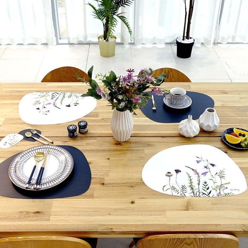 May include: A wooden dining table set with white and navy blue placemats, plates, and cutlery. The table is decorated with a vase of flowers, small white vases, and a plate of fruit. The placemats have floral designs.