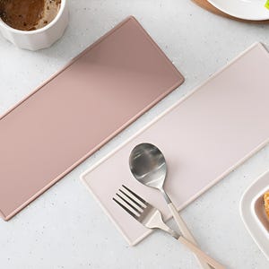 May include: Two rectangular silicone placemats, one in a light pink colour and the other in a darker pink colour. The placemats are on a white surface with a fork and spoon on top of the light pink placemat.