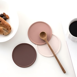 May include: Three round silicone coasters in shades of brown, pink, and beige. A wooden spoon is placed on top of the coasters.