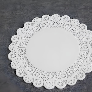 May include: A white paper doily with a decorative, openwork edge. The doily has a circular center and a scalloped border. The intricate design is visible against a gray background. This item is suitable for crafts or table decor.