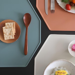 May include: Overhead shot of a table setting with hexagonal plates in teal, peach, and white. A wooden spoon rests on the teal plate next to a small wooden plate with crackers. A plate of food and silverware are on the peach plate.
