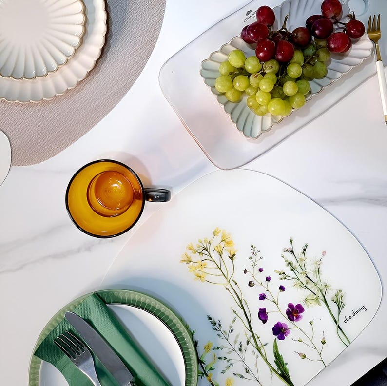 May include: Overhead shot of a table setting with various dishes and food. Includes a white floral placemat, a glass cup with a yellow interior, a plate with a green napkin and silverware, and a rectangular dish with grapes.