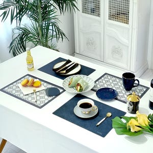 May include: A white table set for a meal with blue placemats, plates, and a coffee cup. The table is decorated with food, cutlery, and a bottle of beverage. A decorative cabinet and a large green plant are in the background.