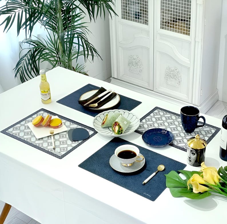 May include: A white table set for a meal with blue placemats, plates, and a coffee cup. The table is decorated with food, cutlery, and a bottle of beverage. A decorative cabinet and a large green plant are in the background.