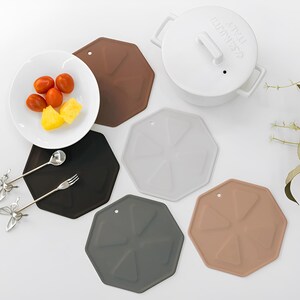 May include: Set of five octagonal silicone hot pads in brown, grey, black, white and beige. The hot pads are designed with a starburst pattern and have a hole in the centre.