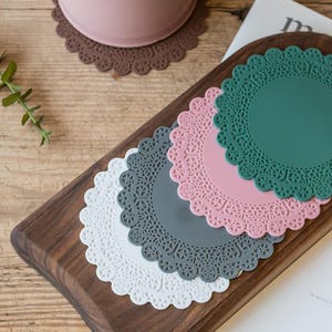 May include: A collection of colorful, round silicone coasters with a lace-like design. The coasters are in shades of white, gray, pink, and green, arranged on a wooden serving tray. A brown coaster and a pink cup are also visible.