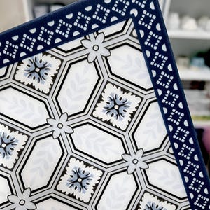 May include: A decorative square plate with a white background featuring a geometric pattern of hexagons, flowers, and leaves in shades of blue, gray, and black. The border is a dark blue with a repeating geometric design.