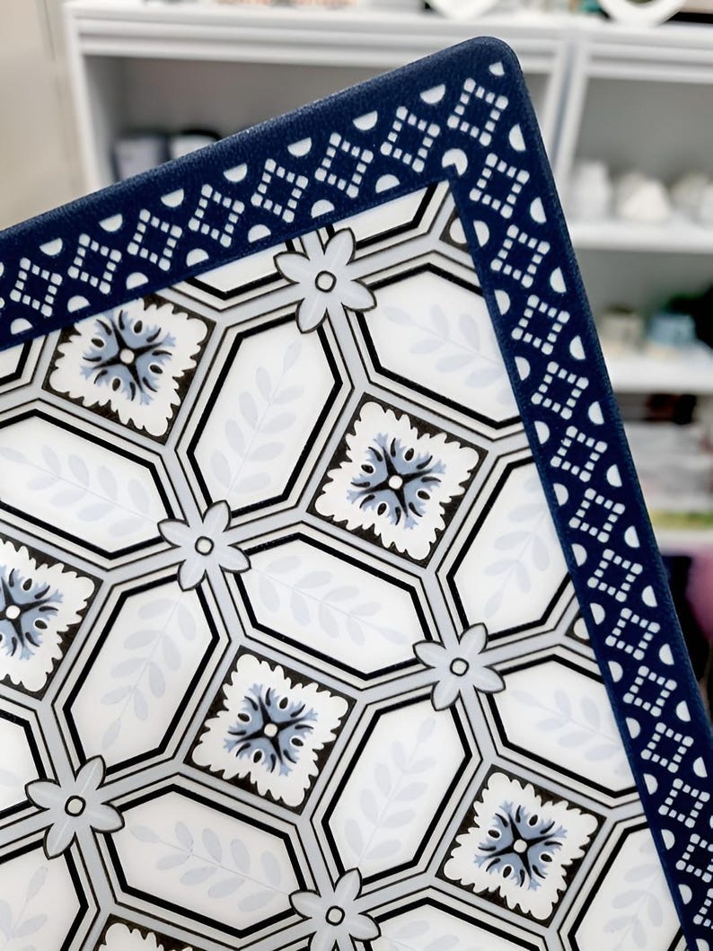 May include: A decorative square plate with a white background featuring a geometric pattern of hexagons, flowers, and leaves in shades of blue, gray, and black. The border is a dark blue with a repeating geometric design.