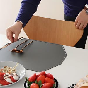 May include: A dining table setting with a gray placemat, copper-colored cutlery, and a plate of food. The table is set with wine glasses and a wooden chair back is visible.